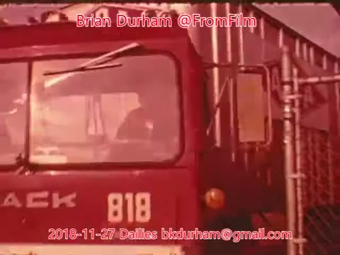 The still depicts a red vehicle, prominently labeled 'PACK 818,' parked near a fence or gate. A figure is visible inside the vehicle, suggesting a moment of waiting or preparing to depart. The overall tone of the image appears warm, likely due to the film's color palette.