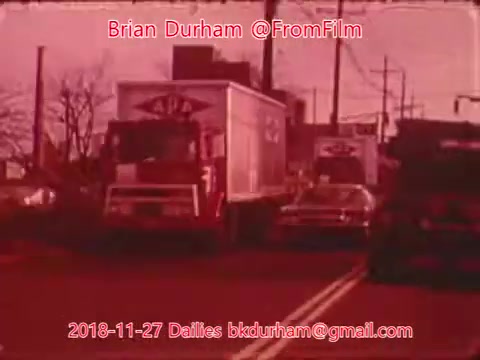 The still features a 16mm film scene depicting a busy street with trucks and cars. The image has a reddish tint, suggesting a nostalgic or vintage aesthetic. There are clear signs of urban infrastructure in the background, such as power lines and buildings. The date and credit information are prominently displayed at the bottom.