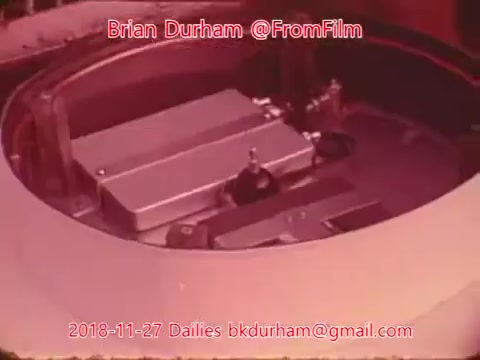 The still depicts a close-up view of a classic film editing setup, likely showing a film reel or editing machine in operation. The surface features intricate machinery, including metal components and parts typical of 16mm film processing. The background appears soft-focused, emphasizing the equipment in the foreground. The image is marked with text indicating the creator and date information.