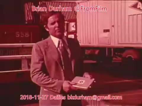 The still features a man in a suit holding a small booklet or folder, standing outdoors near what appears to be trucks or trailers in the background. The scene conveys a professional or promotional context, likely related to a film or advertisement, given the source of the still. The color is warm and vintage, typical of 16mm film.
