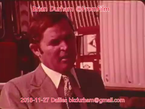 The still features a man in a suit speaking, likely in an interview or presentation setting. The background includes elements that suggest a location related to transportation or industry, possibly a truck or shipping container. The color tone is warm, hinting at a vintage aesthetic typical of 16mm film. Text overlay indicates the date and source of the footage.