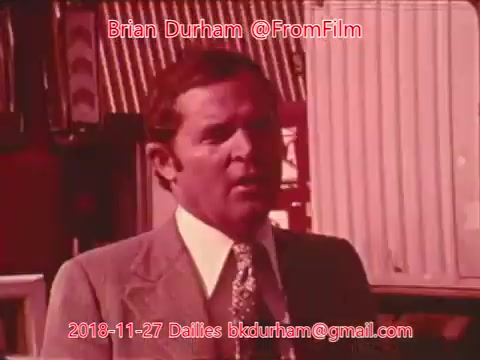 The still features a man in a light-colored suit with a patterned tie, standing against a backdrop of blurred, colorful structures. He appears to be speaking or reacting to something off-camera, with a serious expression. The image is tinted in a reddish hue, contributing to a vintage aesthetic typical of 16mm film. The overlaid text indicates it is a still from the film dailies related to a project titled 'Dristan.'