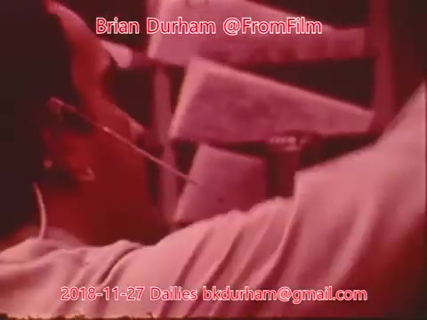 The still appears to depict a person engaged in a task, possibly involving a film-editing process. They seem focused, with tools or materials in the foreground. The image has a vintage aesthetic typical of 16mm film, with a slight color wash that may indicate age or film quality. The scene captures a moment of creativity or production.