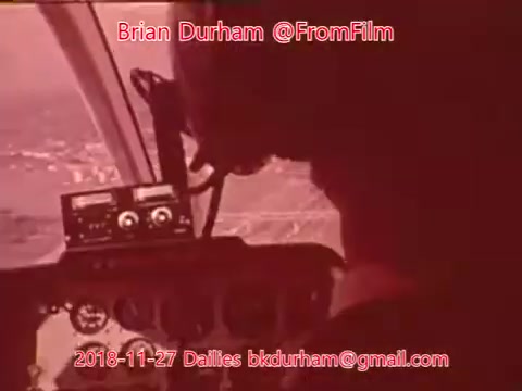 The still features a close-up of an individual operating aircraft controls, with visible gauges and instruments in the cockpit. The scene is tinted in a reddish hue, suggesting an atmospheric or vintage aesthetic. It captures a moment of piloting, likely during a flight, highlighting the focus and engagement of the operator. The date and credits are overlaid at the top and bottom.