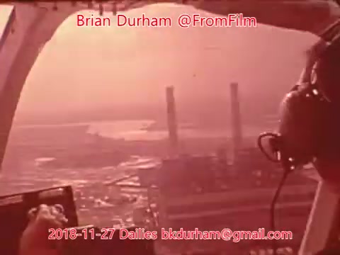 The still depicts a view from a helicopter or aircraft, showing a landscape below with industrial structures, likely power plants, in the background. The environment has a warm, reddish tint typical of 16mm film. A person's hand can be seen operating controls, and they are wearing headphones, indicating they are engaged in aerial observation or filming. The overall atmosphere suggests a documentary or observational context.