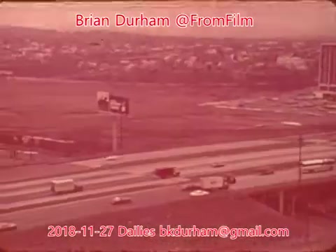 The still captures a highway scene with several vehicles moving along the roadway. In the background, there are buildings and a billboard. The overall tone of the image has a warm red hue, likely due to the film stock used. The date and credit information is also prominently displayed in the image.
