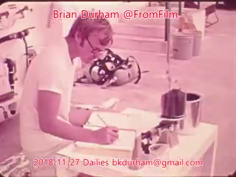 The still shows a person seated at a table, intently focused on writing or drawing. They are dressed in a white outfit and surrounded by various equipment, including what appears to be lab or film-related tools. The background suggests an outdoor or partially enclosed workspace. The image has a vintage 16mm film quality, capturing a moment of concentration in a production environment.