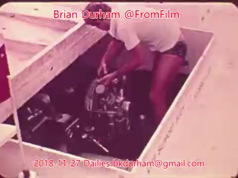 The still shows a man working on an engine compartment, likely a boat or vehicle, in a wooden structure. He appears focused on the task, with his upper body leaning over the engine. The surrounding area is minimal, emphasizing the work being done. The image has a vintage aesthetic, typical of 16mm film.