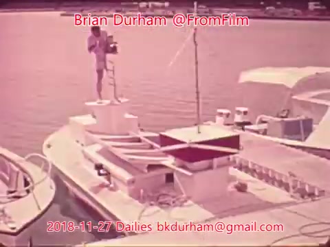 The still depicts a boat on water, possibly in a marina or harbor. A figure stands on the boat, seemingly engaged in an activity, with equipment or tools around. The scene has a vintage aesthetic, likely due to the 16mm film format. The overall tone is serene, capturing a moment on the water.