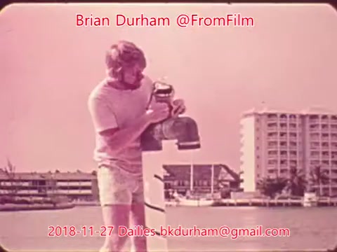 The still depicts a man standing by the water, holding a device or object, likely related to a fishing context. He is wearing light-colored shorts and a shirt, and there are buildings and boats visible in the background, suggesting a coastal or marina setting. The image has a retro aesthetic typical of 16mm film from the era.