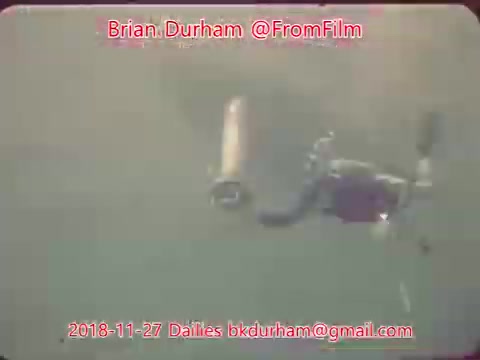 The still features an underwater scene with a diver holding what appears to be a camera or equipment. The water has a murky appearance, suggesting a natural setting. The image is likely part of film dailies, capturing a moment from underwater filming activities.