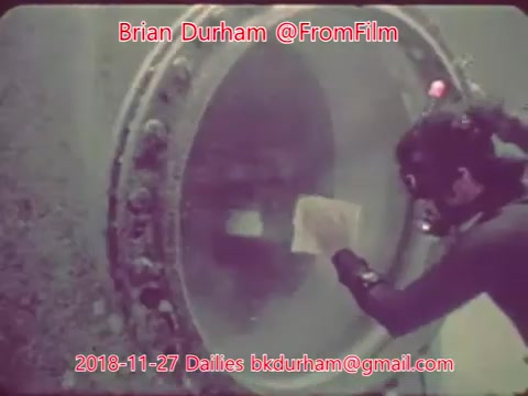 The still features a diver working near a large, circular window, likely part of an underwater habitat or structure. The diver appears to be holding a piece of paper or material while interacting with the environment. The underwater scene has a muted color palette, hints of light filtering through the water, and showcases a sense of exploration and curiosity.