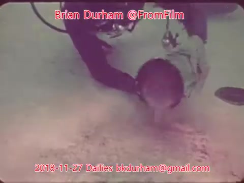 The still likely shows a scene from underwater filming, featuring a diver interacting with a submerged object or possibly assisting another individual. The image is tinted and has a vintage feel typical of 16mm film captures. The context suggests it may be part of a project or exploration related to underwater environments or activities.