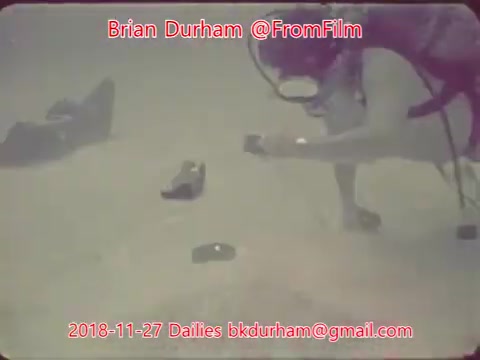 The still depicts a diver underwater, focused on examining an object on the ocean floor. The scene is characterized by a muted color palette typical of 16mm film, with a sense of calmness and curiosity. Surrounding the diver are what appear to be shoes or other items scattered on the seabed, hinting at the exploration theme. The timestamp and contact information indicate it’s part of a dailies collection from November 27, 2018.