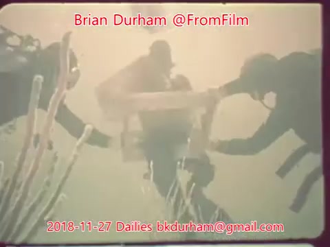 The still appears to capture an underwater scene featuring divers in wet suits. They are engaged in an activity, possibly involving equipment or a scientific task, while surrounded by aquatic flora. The image is framed with a vintage aesthetic, typical of 16mm film.