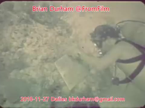 The still features an underwater scene with a diver examining an object on the ocean floor. The diver is equipped with standard scuba gear, and the environment appears rocky with aquatic life possibly visible in the blurred background. The image carries a watermark with the contact information of Brian Durham, indicating it’s part of film dailies from November 27, 2018.