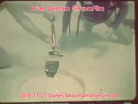 The 16mm still depicts two individuals working near a circular object in sandy terrain. One person appears to be using a tool or device, possibly for measurement or excavation, while the other is positioned nearby, observing or assisting. The setting suggests a focus on an outdoor or experimental activity.