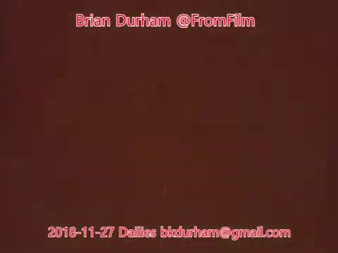 The still features a simple, textured background in a reddish hue. Superimposed text includes the name 'Brian Durham,' the handle '@FromFilm,' and the date '2018-11-27 Dailies,' along with an email address. The composition suggests a focus on film-related work or daily footage updates.