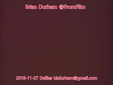 The still from '@FromFilm Dailies' dated November 27, 2018, features a textured background in a reddish hue. The text overlays include the name Brian Durham and contact information, suggesting it's related to film dailies or production notes. The visual aspect emphasizes color and text rather than imagery or action.