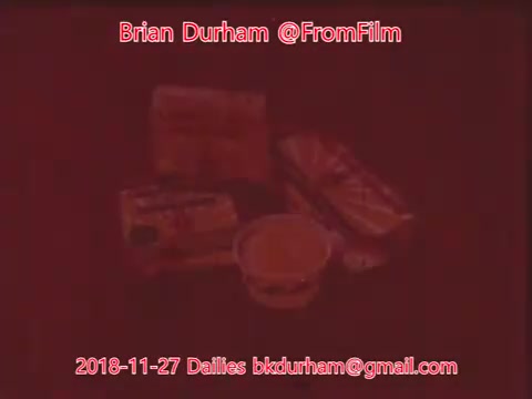 The still features a collection of packaged food items arranged on a surface. The image appears to have a reddish tint, obscuring some details, but you can see items like a cup and several snack packages. Overlaid text identifies the date, the creator, and an email address.