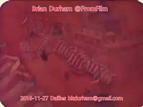 The 16mm still appears to depict a vibrant, possibly surreal scene with strong red hues. It includes abstract shapes and patterns, which may suggest a carnival or festive atmosphere. The image is marked with details about its creator, date, and email, indicating a focus on film dailies from a specific production or project. The overall composition seems to emphasize color and form over clear representation.