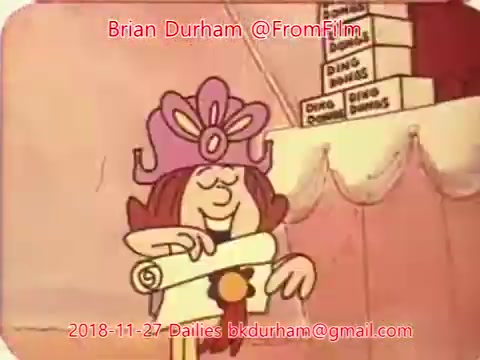 The still features a joyful animated character wearing a large pink hat adorned with a flower. They are holding a scroll and smiling, suggesting excitement or anticipation. In the background, there are stacks of boxes labeled 'DING DINGS.' The overall ambiance is playful and whimsical, characteristic of a lighthearted animated scene.