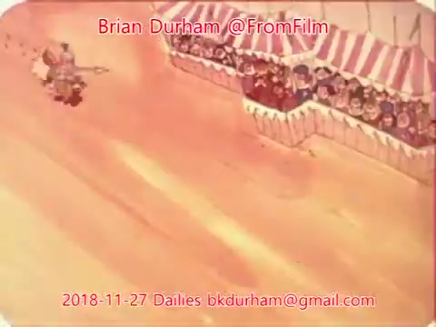 The still appears to depict an animated scene featuring a character, possibly a performer, in a dynamic pose. In the background, a crowd watches from behind striped tents, suggesting a carnival or fair atmosphere. The overall color palette is warm, creating a lively and engaging mood. The image includes text identifying the date and source.