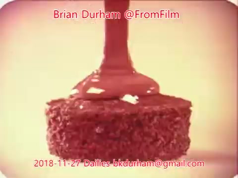 The still features a close-up of a piece of cake being drizzled with chocolate frosting. The texture of the cake is visible, with the chocolate cascading elegantly on top. The background has a soft focus, emphasizing the cake as the main subject. The image appears warm and inviting, typical of nostalgic film aesthetics.
