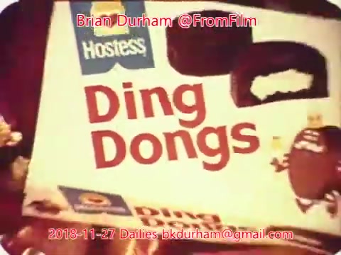 The still features a box of Hostess Ding Dongs, prominently displaying the product name in bold letters. The image has a nostalgic, vintage quality, typical of 16mm film, with a warm color palette. The background appears cluttered, hinting at a casual setting, possibly a kitchen or a living room, evoking a sense of familiarity and comfort. The date and source information is overlaid at the bottom.