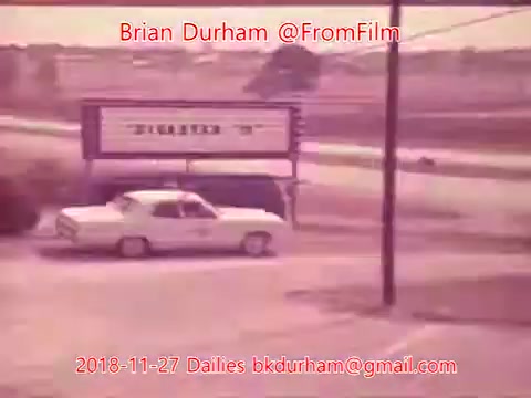 The 16mm still from the '@FromFilm Dailies November 27, 2018' depicts a white car on a road near a sign with the word 'Studio.' The image has a vintage aesthetic, suggesting a retro time period. The surrounding landscape appears somewhat rural, with a clear sky and minimal traffic. The photo captures a moment frozen in time, emphasizing the simplicity of the scene.