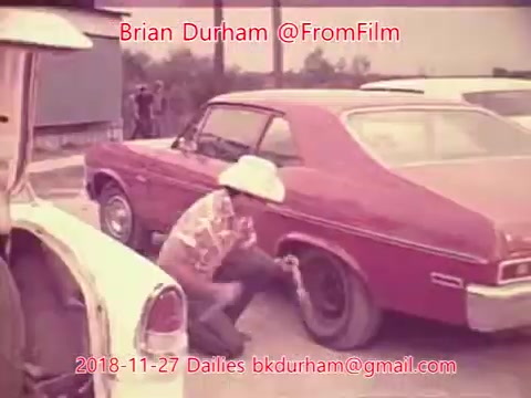 The still features a man wearing a large white hat, crouched by a red car, possibly fixing or inspecting a tire. In the background, there are other vehicles and a few individuals, suggesting an outdoor setting, likely a roadside or parking area. The overall tone appears nostalgic, typical of 16mm film aesthetics. Text overlaid includes the date, context, and contact information.
