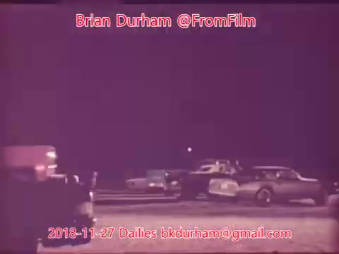 The still features a nighttime scene with several parked cars illuminated under dim light. An ambulance or emergency vehicle is positioned on the left side, while other vehicles are scattered throughout the background. The atmosphere is quiet and somewhat nostalgic, hinting at a setting that might suggest a gathering or an event. The overall tone is muted, reflecting the characteristics of 16mm film.