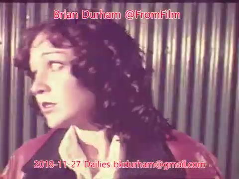 The still features a woman with curly hair, looking off-camera, conveying a sense of concern or curiosity. She is dressed in a jacket over a light shirt. The background shows vertical metal panels, which adds an industrial feel to the scene. The image has a vintage quality characteristic of 16mm film.