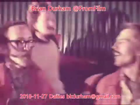 The still appears to show a scene with two men, possibly engaged in a lively conversation or performance, both wearing red outfits. One man has glasses and a mustache, while the other has a more distinctive facial expression. The setting seems illuminated with warm lighting, and there is a sense of camaraderie or fun in the moment captured. The image includes text that indicates it is a still from a film dailies project by Brian Durham.