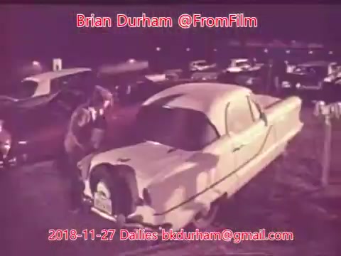 The still features a vintage white car parked among other vehicles in a dimly lit setting. A person is seen interacting with the car, possibly examining it. The color tones have a nostalgic quality, reminiscent of old film. The date and contact information prominently displayed suggests it's part of a film daily collection.