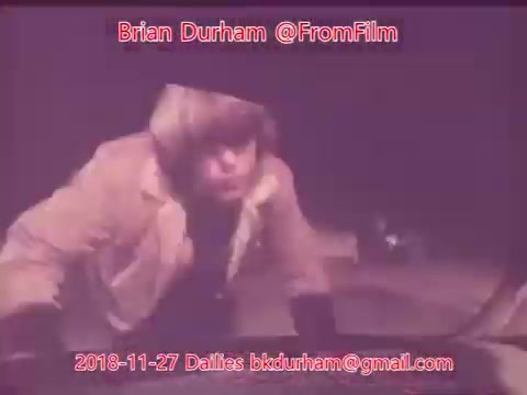 The still features a young man, possibly in an outdoor or low-light setting, wearing a light-colored jacket. He appears to be engaged in some activity, with a focused expression. The image is tinted with a warm hue, adding a vintage feel typical of 16mm film. Text overlays indicate the creator's name and date related to the film dailies.