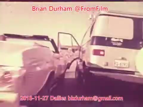 The still from '@FromFilm Dailies November 27, 2018' depicts a traffic scene featuring a vintage car with an open driver's door, seemingly parked or stopped next to a van. The image has a retro aesthetic, indicative of a 16mm film, and includes text credits. The composition suggests a moment of pause or transition within a busy environment.