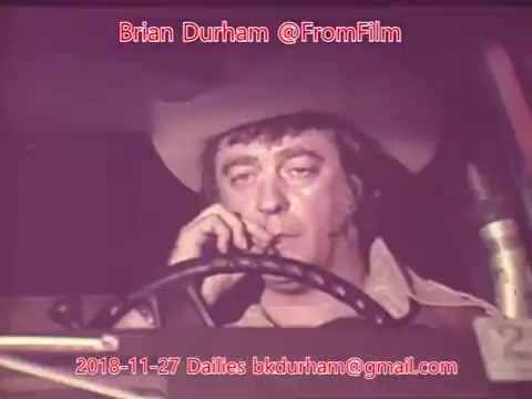 The still features a man wearing a cowboy hat, sitting behind the wheel of a vehicle. He appears to be smoking, with a serious expression on his face. The image has a retro aesthetic typical of 16mm film, showcasing a slightly faded color palette. Text overlays include the name 'Brian Durham,' the handle '@FromFilm,' and a date, indicating its context within film dailies from November 27, 2018.