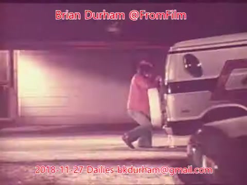 The still captures a scene from a film, featuring a person in a pink shirt and blue jeans, seemingly working on or interacting with a white van. The background suggests an urban setting, with a garage visible. The image has a vintage 16mm film quality, contributing to a nostalgic atmosphere.