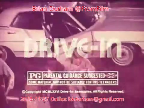 The still features a retro drive-in movie set-up, with a prominent title 'DRIVE-IN' displayed. It includes a 'PG' parental guidance label, indicating the content may not be suitable for younger viewers. The visual style is reminiscent of classic cinema, with a muted color palette typical of 16mm film. People can be seen outside of vehicles, likely engaging with the drive-in experience. The date and name associated with the film dailies, November 27, 2018, and contact information are also present.