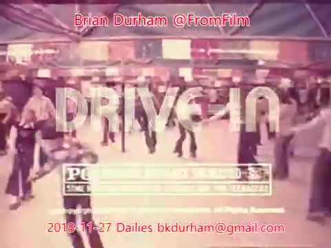 The 16mm still from '@FromFilm Dailies November 27, 2018' titled 'ffn035-dristan-dailies' depicts a lively scene at a drive-in event. The image features people dancing and socializing in a vibrant atmosphere, possibly in a retro or nostalgic setting. The word 'DRIVE-IN' is prominently displayed, suggesting a focus on classic drive-in culture, capturing the essence of communal entertainment.