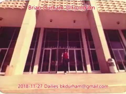 The still appears to be a scene captured from a 16mm film, showing a person standing outside a modern building entrance with large glass doors. The architecture features vertical lines and a textured facade. The individual is dressed in red, standing under an awning, suggesting an inviting yet formal environment. The image likely has a nostalgic quality, characteristic of film dailies from the late 2010s.