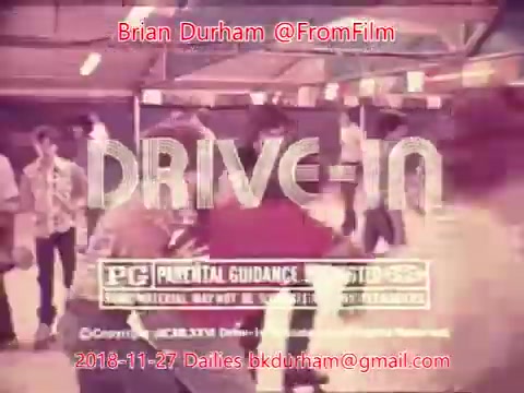 The still depicts a vibrant drive-in scene, showcasing a mix of people engaged in various activities. An overlay with the title 'DRIVE-IN' is prominently featured, along with PG rating information. The image has a nostalgic feel, reminiscent of classic cinema, with a soft, aged appearance typical of 16mm film.
