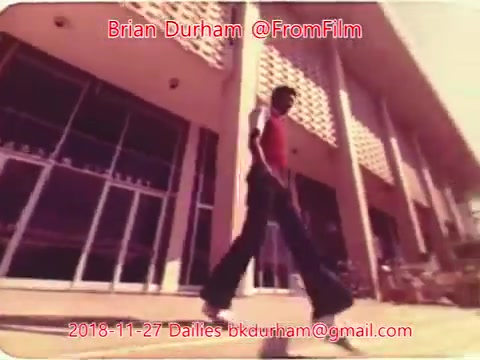 The still captures a figure walking in front of a modern building, characterized by large windows and a geometric façade. The individual is dressed in a vibrant outfit, prominently featuring a red top and flared black pants, conveying a retro style. The lighting appears warm, suggesting a sunny day. The image has a nostalgic quality typical of 16mm film, with a slight graininess enhancing its vintage feel.