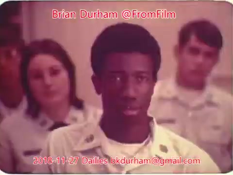 The still features a young Black man in the foreground, with a serious expression, surrounded by a group of individuals in uniform, likely in a classroom or instructional setting. The background has two individuals, including a woman and another man, both appearing focused on the speaker or topic at hand. The overall tone suggests an educational or training environment. The image carries a vintage aesthetic, characteristic of 16mm film.