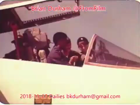 The still appears to show two individuals in the cockpit of an aircraft, engaged in conversation. They seem to be wearing aviation-related attire, and the cockpit is well-equipped with instruments visible. The image has a vintage quality, typical of 16mm film, and includes text indicating the date and origin of the footage. Overall, it captures a moment of interaction in an aviation setting.