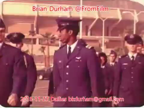 The still features a group of military personnel in uniform, walking in formation. The setting appears to be an outdoor location, possibly near a military base or training facility, with a structure visible in the background. The focus is on a man in the front, who stands out due to his posture and expression. The overall tone is reminiscent of mid-20th century military life.