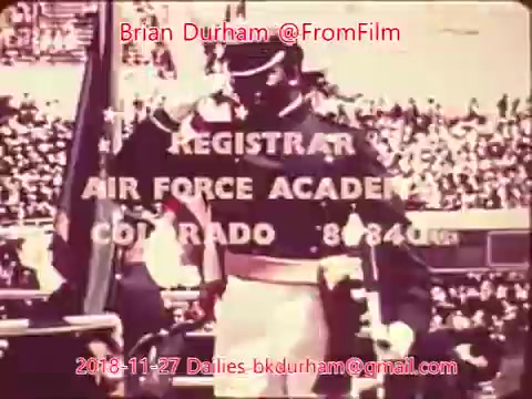 The 16mm still displays a military figure saluting, likely at a ceremonial event, with a large crowd in the background. Text overlays indicate the 'Registrar' of the 'Air Force Academy' in Colorado, along with a contact email and the date of the film dailies. The image has a vintage quality typical of 16mm film.