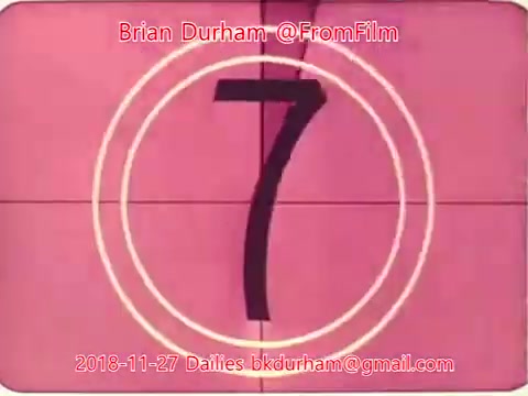 The image shows a still frame from a 16mm film, featuring a countdown timer with the number '7' prominently displayed in a bold, vintage style. The background is a soft pink color, and there are circular frames surrounding the number, typical of classic film countdowns. The top and bottom of the image include text indicating the creator (Brian Durham @FromFilm) and the date (November 27, 2018), along with an email address. The overall aesthetic reflects a retro film look.