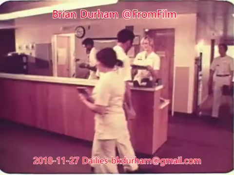 The still captures a scene set in a medical or hospital environment. Several staff members in uniforms are engaged in activities at a reception area. A clock is visible on the wall, indicating the setting's busy nature. The overall atmosphere suggests a typical day in a healthcare facility, with workers attending to tasks and interacting with one another. The color palette appears warm, reflecting the vintage 16mm film aesthetic.